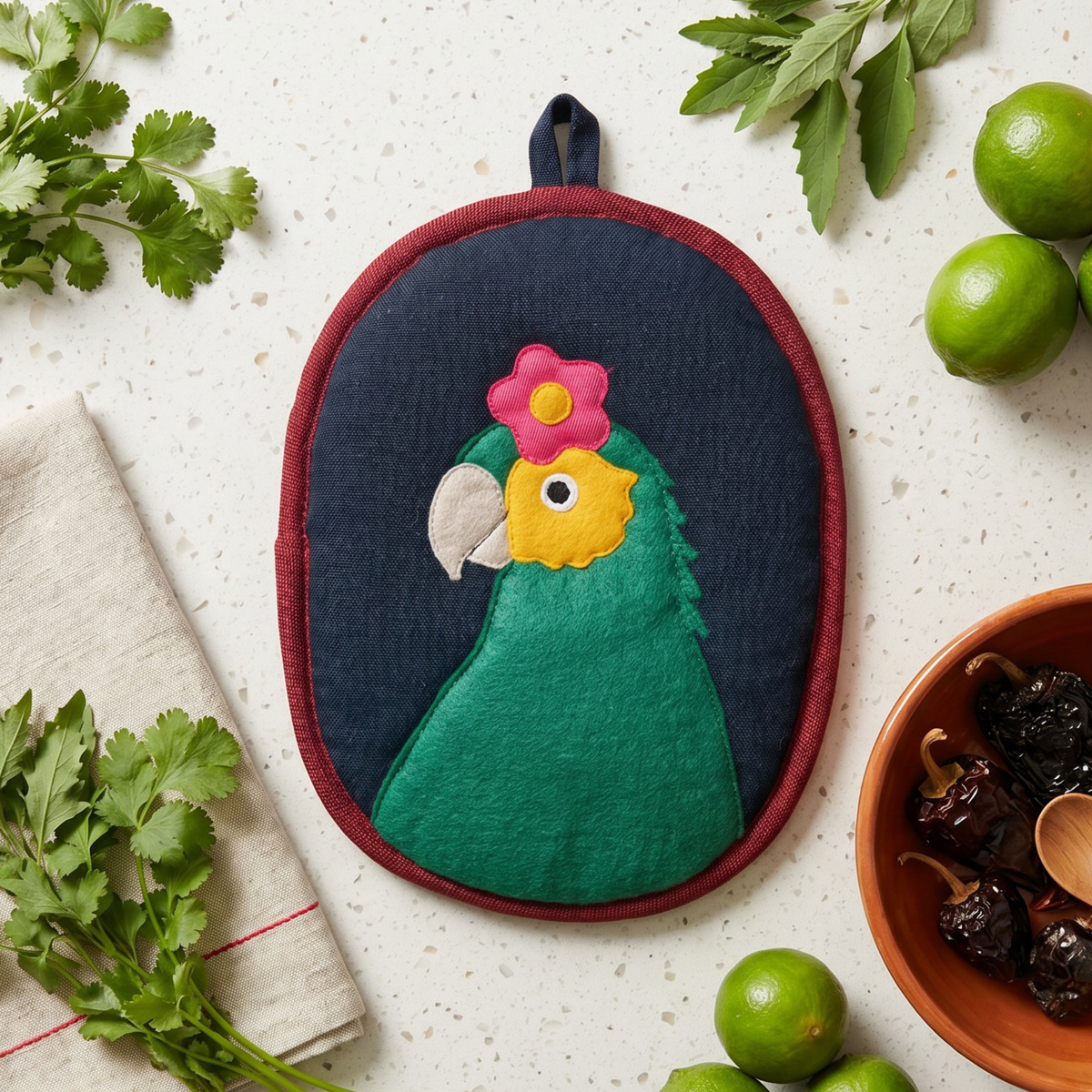 An oval potholder with a colorful parrot design, styled in a flat-lay on a speckled white countertop with limes, cilantro, and a bowl of dried chilies.