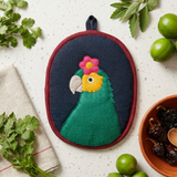 An oval potholder with a colorful parrot design, styled in a flat-lay on a speckled white countertop with limes, cilantro, and a bowl of dried chilies.