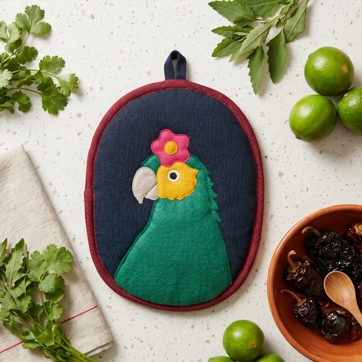 An oval fabric potholder featuring an embroidered green parrot with a pink flower on its head, set against a dark navy background with a red border. The potholder is styled in a flat-lay arrangement on a speckled white countertop with limes, cilantro, a bowl of dried chilies, and a napkin.