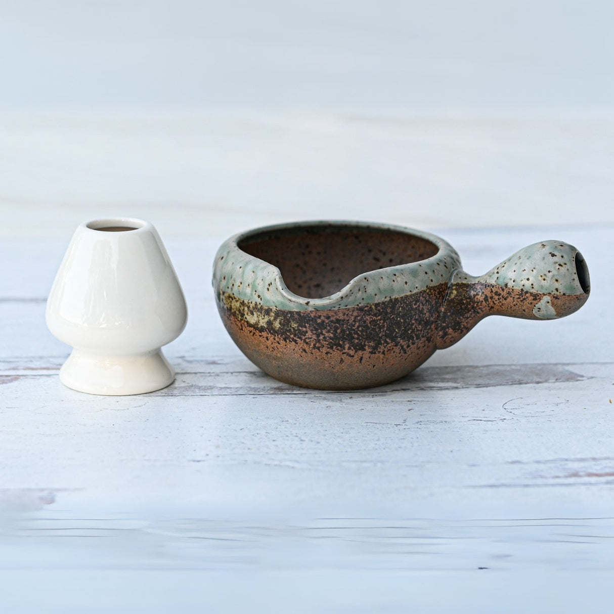 A small white vase and a Brown Ceramic Matcha Bowl with Spout, evoking the essence of an organic Japanese tea ceremony, rest on a textured surface.