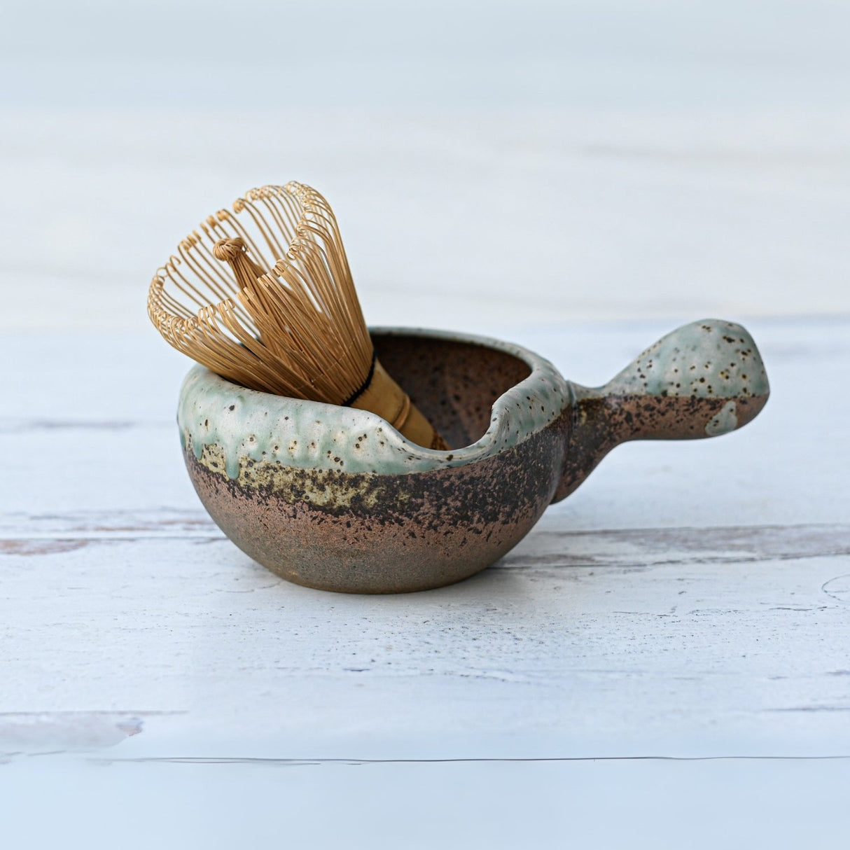 The Brown Ceramic Matcha Bowl with Spout cradles a bamboo whisk on light wood, part of an elegant set including a whisk holder and organic Japanese matcha for a refined tea ceremony experience.