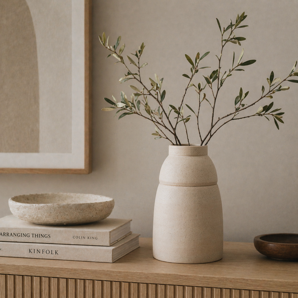 Beige vase with green branches on a wooden surface with books and a framed picture in the background.
