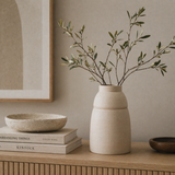 Beige vase with green branches on a wooden surface with books and a framed picture in the background.