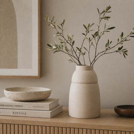 Beige vase with green branches on a wooden surface with books and a framed picture in the background.