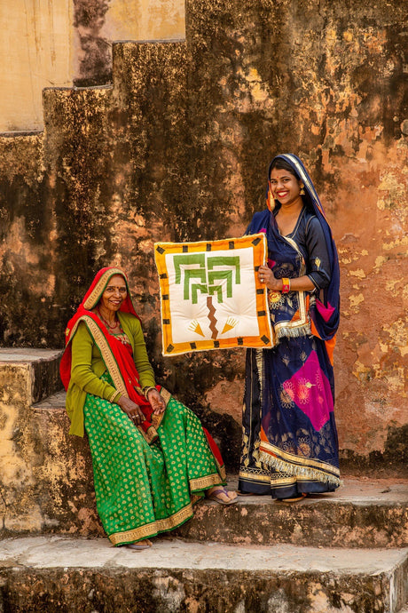 Two women sitting on stone steps holding a colorful fabric in front of an old wall.