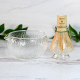 The Textured Spouted Glass Matcha Bowl Pattern 2 and Bamboo Matcha Whisk with a Whisk Holder Set sit elegantly on a white surface, accented by green tea leaves in the background.