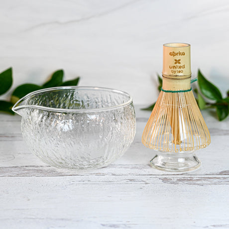 The Textured Spouted Glass Matcha Bowl Pattern 2 and Bamboo Matcha Whisk with a Whisk Holder Set sit elegantly on a white surface, accented by green tea leaves in the background.