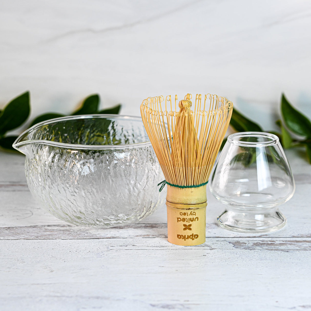 The Textured Spouted Glass Matcha Bowl Pattern 2, a bamboo matcha whisk, and a small glass cup rest elegantly on a white surface with green leaves providing a serene backdrop.