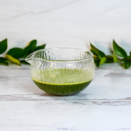 A Textured Spouted Glass Matcha Bowl Pattern 2 with green tea is on a white wooden surface, next to a bamboo matcha whisk, with green leaves in the background.
