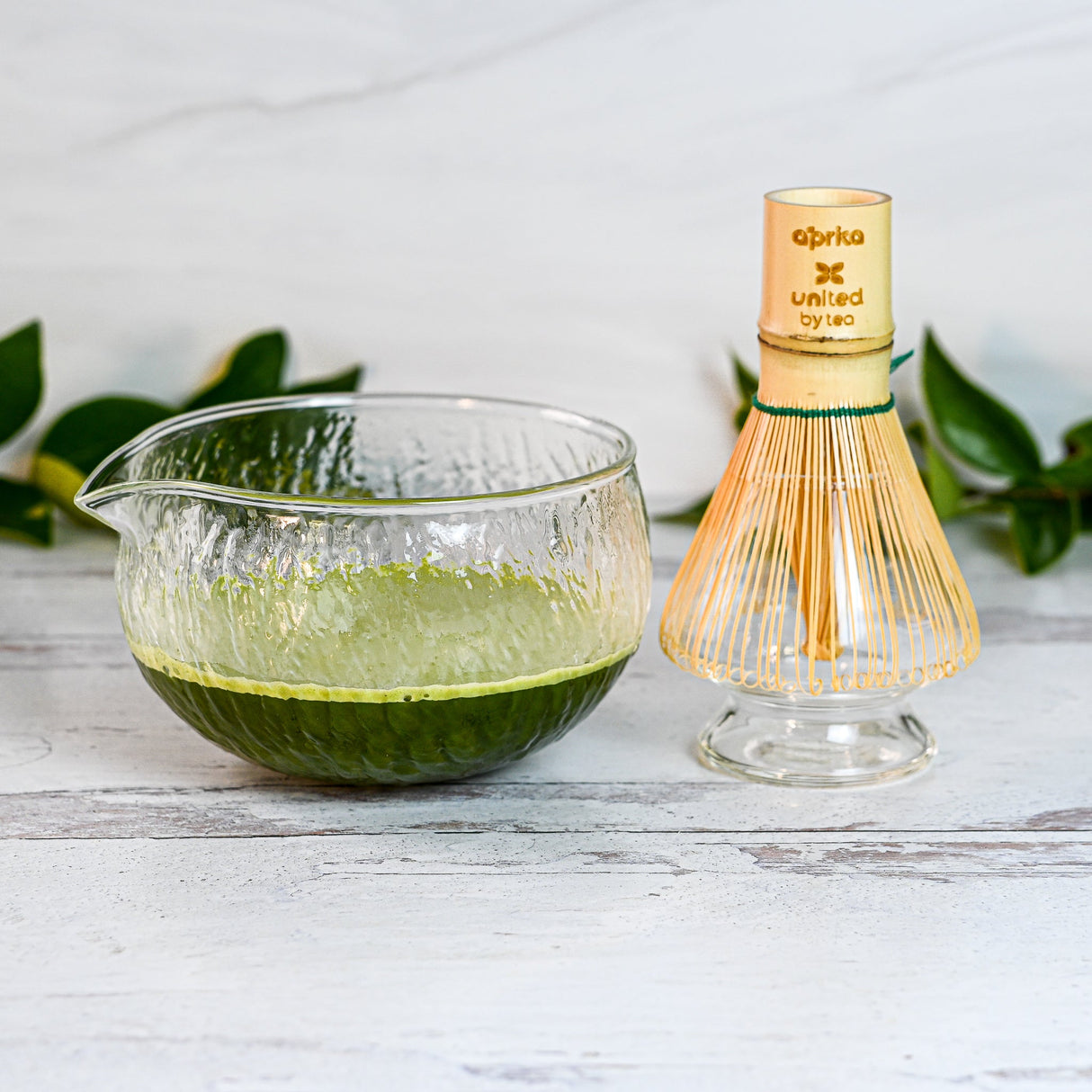 A Textured Spouted Glass Matcha Bowl Pattern 2, filled with vibrant green matcha, sits beside a Bamboo Matcha Whisk and Whisk Holder Set on a light wooden surface, with green leaves in the backdrop for serenity.