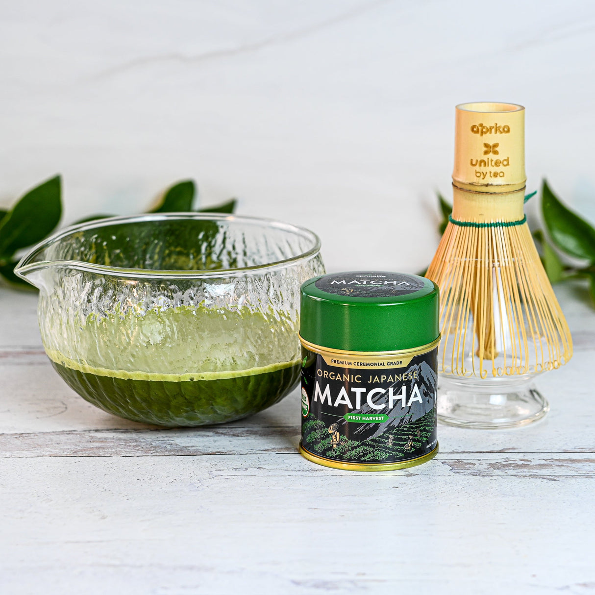 The Textured Spouted Glass Matcha Bowl Pattern 2, filled with vibrant green matcha tea, is arranged on a pristine white surface alongside a sleek matcha tin and a bamboo whisk.