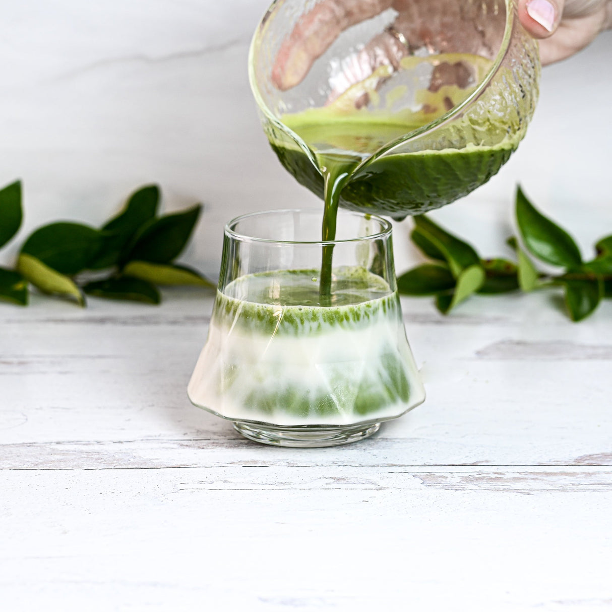 A vibrant green liquid flows into a glass of milk, surrounded by lush leaves. Nearby, a bamboo matcha whisk and the Textured Spouted Glass Matcha Bowl Pattern 2 await their turn in this tranquil scene.