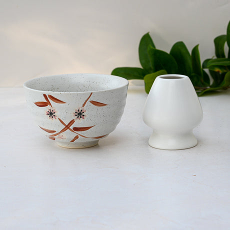 A Bamboo Matcha Bowl featuring a floral design rests beside a small white vase on a light surface, highlighted by green leaves in the background.