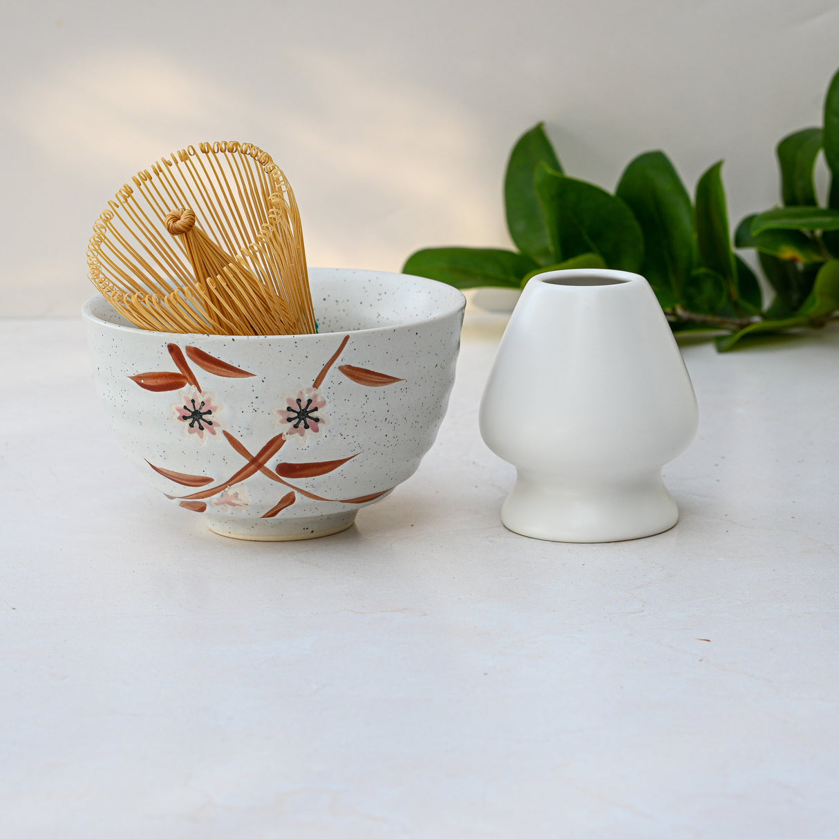 The Bamboo Matcha Bowl, Whisk, and Holder Set sit on a light surface surrounded by green leaves in the background.
