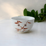 Bamboo Matcha Bowl with floral design on a light background, complemented by a Bamboo Matcha Whisk and scattered green leaves.