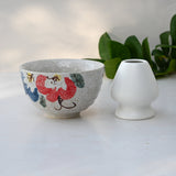 The Red and Blue Flowers Matcha Bowl, part of a matcha set with a bamboo whisk and holder, sits gracefully beside a small white vase on a light surface with green leaves, highlighting the elegance of Minoyaki pottery techniques.
