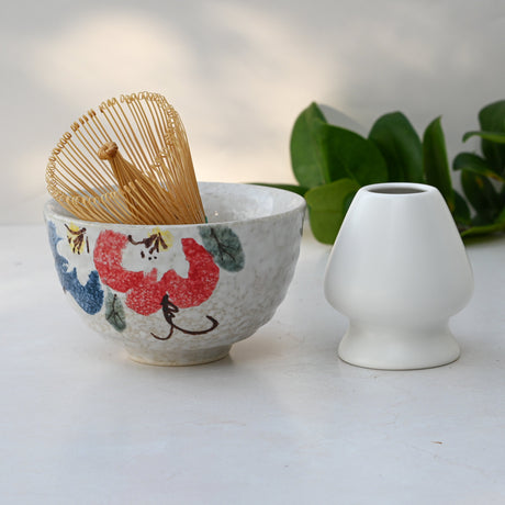 The Red and Blue Flowers Matcha Bowl, with a bamboo whisk and holder inside, sits beside a white vase with green leaves in the background, showcasing Minoyaki pottery techniques.