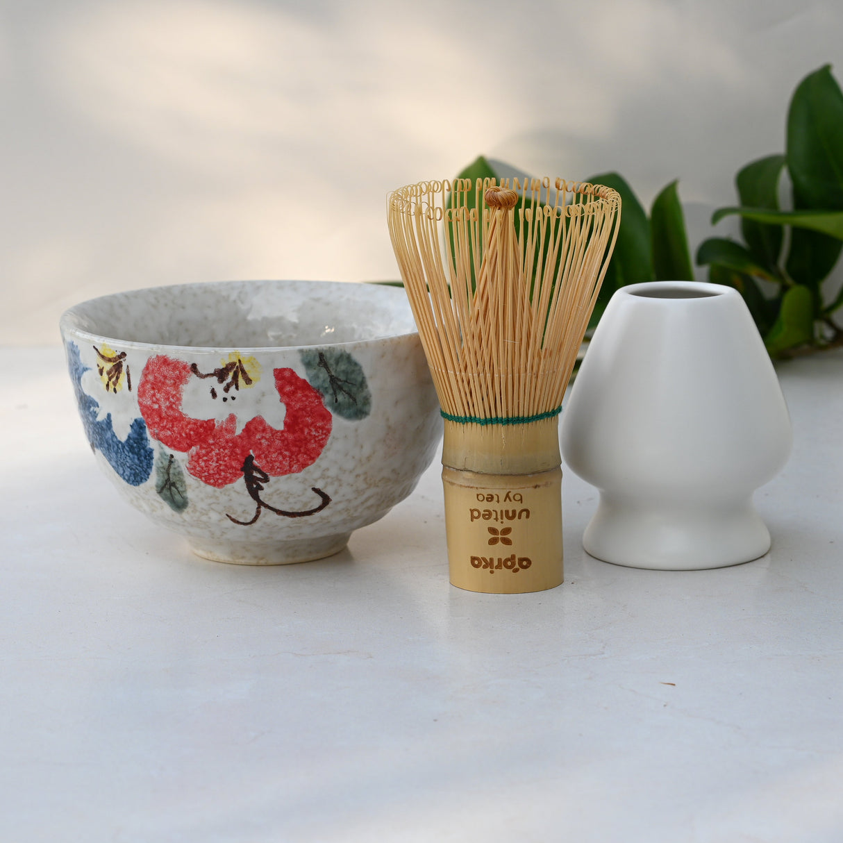 On a light surface with green leaves, the Red and Blue Flowers Matcha Bowl, Bamboo Matcha Whisk, and Whisk Holder Set showcases Minoyaki pottery techniques.