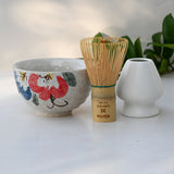 On a light surface with green leaves, the Red and Blue Flowers Matcha Bowl, Bamboo Matcha Whisk, and Whisk Holder Set showcases Minoyaki pottery techniques.