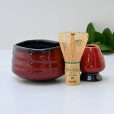 A Red Matcha Bowl, crafted with exquisite Minoyaki pottery and accompanied by a matcha whisk, is displayed elegantly on a white surface with green leaves in the background.