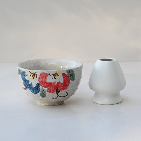 A Red and Blue Flowers Matcha Bowl, crafted with Minoyaki pottery techniques, displays colorful floral designs alongside a small white vase on a light surface.