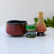 A red matcha bowl, green tea tin, and bamboo whisk rest on a white surface with lush greenery in the background, capturing the essence of the Red Matcha Bowl, Bamboo Matcha Whisk, Whisk Holder and Organic Japanese Matcha Set.