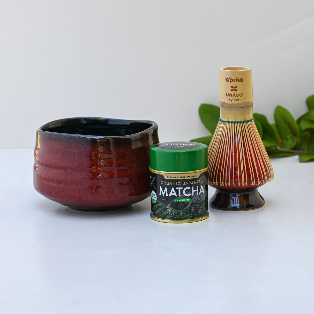 A red matcha bowl, green tea tin, and bamboo whisk rest on a white surface with lush greenery in the background, capturing the essence of the Red Matcha Bowl, Bamboo Matcha Whisk, Whisk Holder and Organic Japanese Matcha Set.