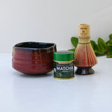 A red matcha bowl, green tea tin, and bamboo whisk rest on a white surface with lush greenery in the background, capturing the essence of the Red Matcha Bowl, Bamboo Matcha Whisk, Whisk Holder and Organic Japanese Matcha Set.