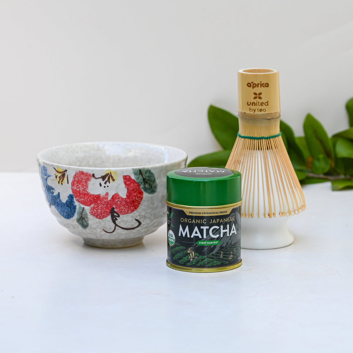 The Red and Blue Flowers Matcha Bowl, complemented by a bamboo matcha whisk and whisk holder set, beautifully arranged on a white surface with green leaves softly framing the scene.