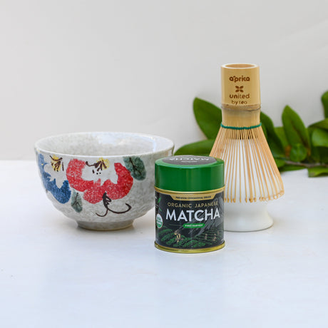 The Red and Blue Flowers Matcha Bowl, complemented by a bamboo matcha whisk and whisk holder set, beautifully arranged on a white surface with green leaves softly framing the scene.