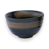 The Dark Brown Glaze Ceramic Matcha Bowl is crafted from black ceramic using Minoyaki pottery techniques, featuring a glossy, uneven surface with a brown stripe around the middle on a black background.