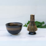 The Dark Brown Glaze Ceramic Matcha Bowl and Bamboo Matcha Whisk from the Organic Japanese Matcha Set rest on a white surface, with green leaves softly blurring in the background.