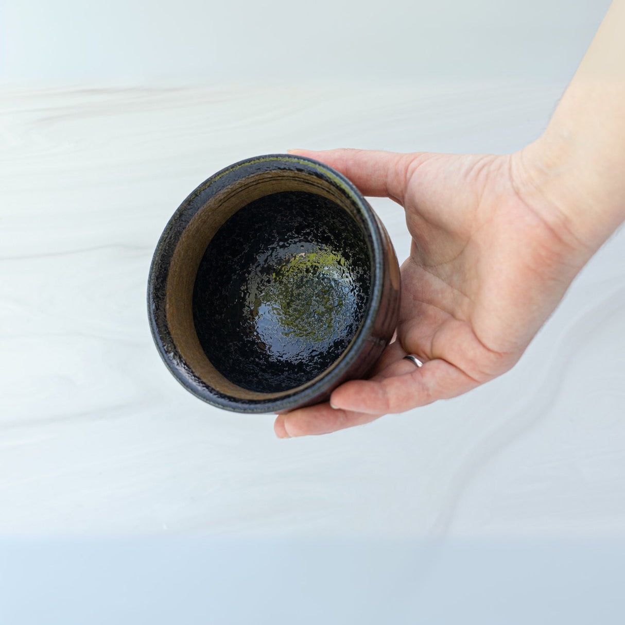 A hand holds an empty Dark Brown Glaze Ceramic Matcha Bowl from the Organic Japanese Matcha Set, crafted with Minoyaki pottery techniques against a marble-like background.