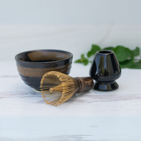 The Dark Brown Glaze Ceramic Matcha Bowl Set includes a bamboo whisk and holder, arranged elegantly on a white surface.