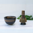 The Dark Brown Glaze Ceramic Matcha Bowl and Bamboo Matcha Whisk from the Organic Japanese Matcha Set rest on a white surface, with green leaves softly blurring in the background.