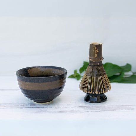 The Dark Brown Glaze Ceramic Matcha Bowl and Bamboo Matcha Whisk from the Organic Japanese Matcha Set rest on a white surface, with green leaves softly blurring in the background.