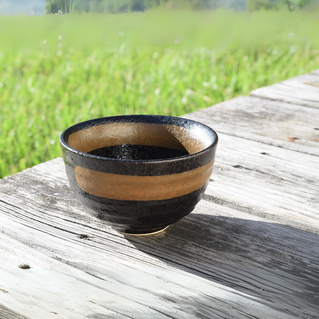 The Dark Brown Glaze Ceramic Matcha Bowl, inspired by traditional Minoyaki pottery, rests on a rustic wooden table with a blurred grassy background.