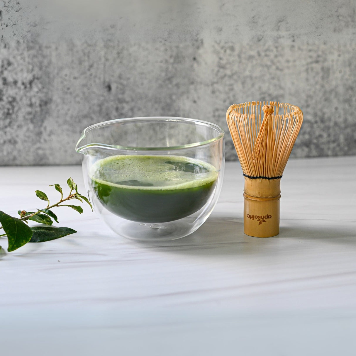 On a white surface sits a vibrant green matcha tea within a Double Sided (Double Wall) Glass Matcha Bowl with Spout, accompanied by a bamboo whisk and scattered leaves.