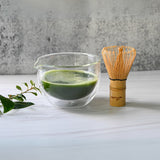 On a white surface sits a vibrant green matcha tea within a Double Sided (Double Wall) Glass Matcha Bowl with Spout, accompanied by a bamboo whisk and scattered leaves.
