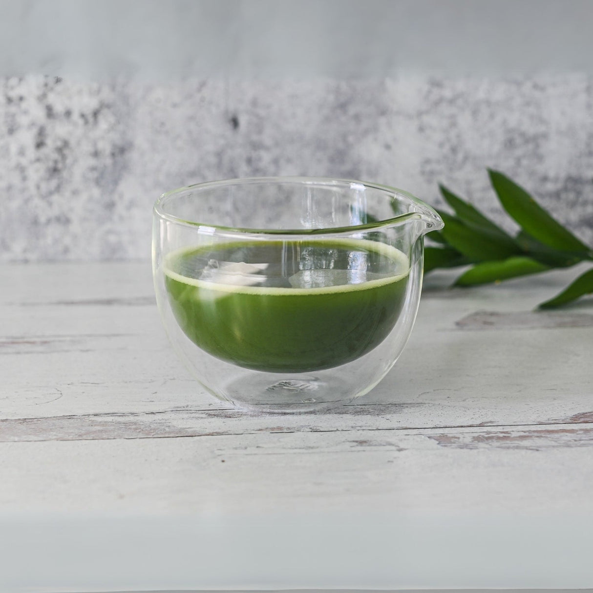 A Double Sided Glass Matcha Bowl with Spout, filled with vibrant green tea, sits on a light wooden surface, complemented by fresh green leaves. Complete your serene tea ritual with this set that includes a Bamboo Matcha Whisk and Whisk Holder.