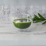 A Double Sided Glass Matcha Bowl with Spout, filled with vibrant green tea, sits on a light wooden surface, complemented by fresh green leaves. Complete your serene tea ritual with this set that includes a Bamboo Matcha Whisk and Whisk Holder.