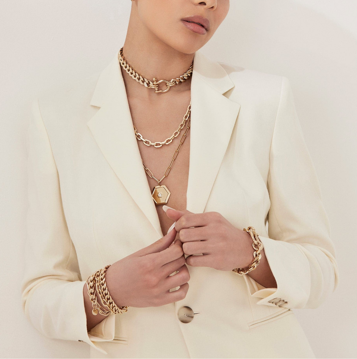 Person wearing a cream blazer with gold jewelry including necklaces, bracelets, and rings.
