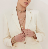 Person wearing a cream blazer with gold jewelry including necklaces, bracelets, and rings.