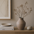 Ceramic vase with dried branches on a wooden surface, with books and a bowl in the background.