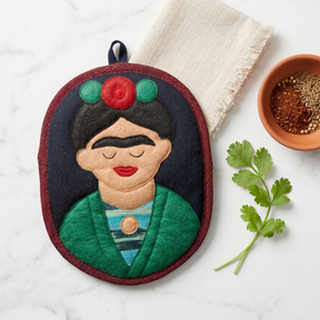 An oval fabric potholder featuring an embroidered Frida Kahlo-style portrait, styled on a white marble surface with a folded cream cloth, a small bowl of spices, and cilantro.