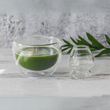 A Double Sided Glass Matcha Bowl with vibrant green liquid sits beside an empty glass on a white wooden surface, accented by a green leaf. This serene scene is perfect for using the Bamboo Matcha Whisk and Holder from the Double Wall Matcha Tea Set.