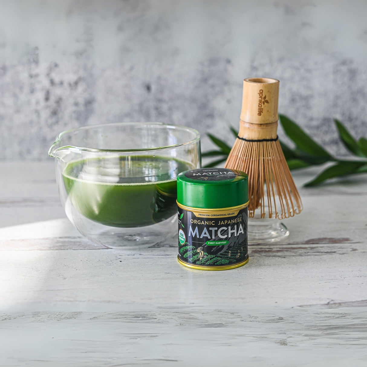 A Japanese Double Sided Glass Matcha Bowl with a spout, a tin of vibrant green tea powder, and a delicate bamboo whisk elegantly rest on a white wooden surface.