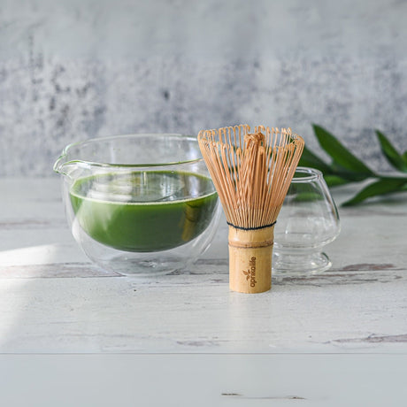 Explore the elegance of a Double Sided Glass Matcha Bowl with Spout, paired with a Bamboo Matcha Whisk and Whisk Holder Set. Experience vibrant leaves and an empty glass cup on a light surface, inviting you to indulge in the art of matcha preparation.