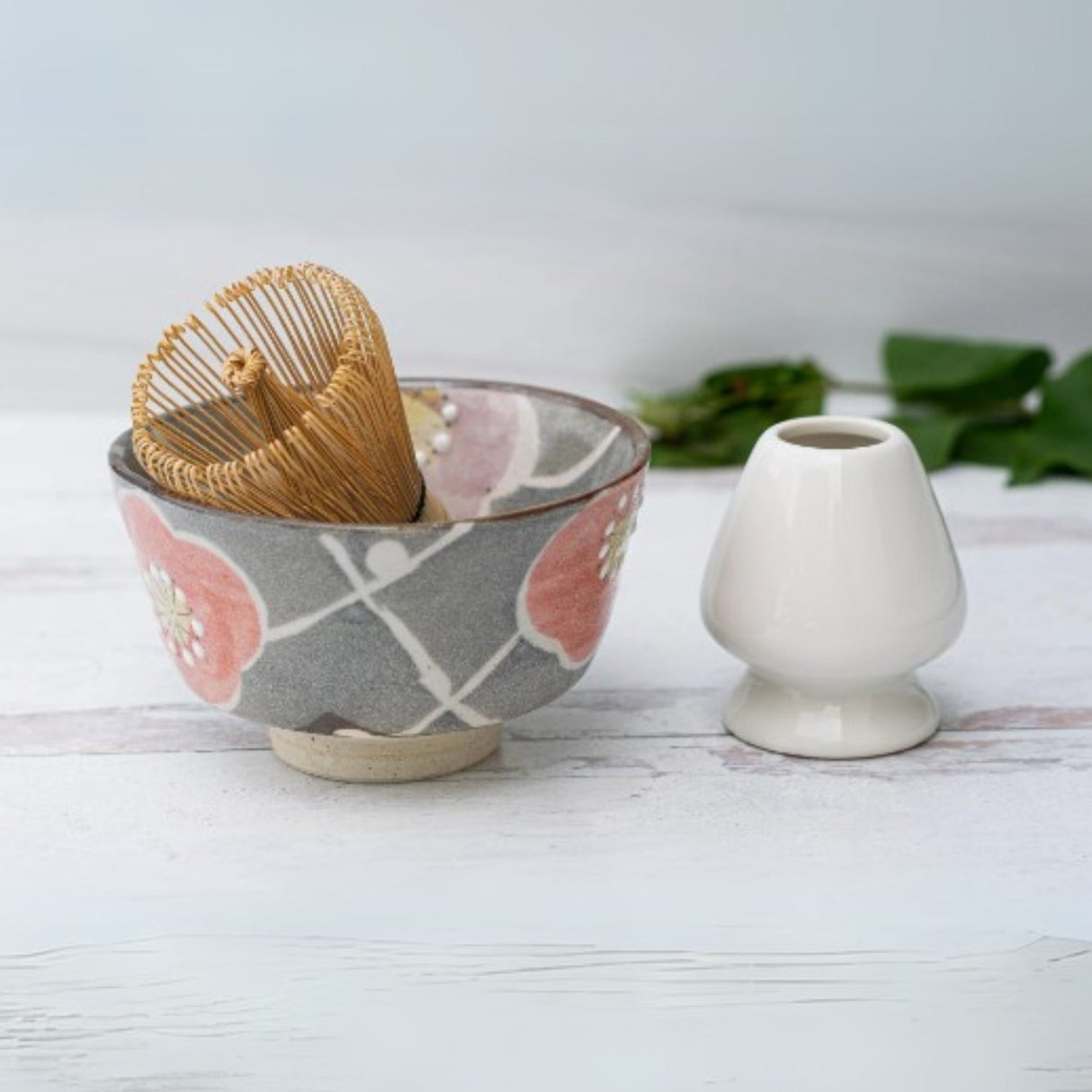 The Gray with Flowers Ceramic Matcha Bowl, Bamboo Matcha Whisk, and Whisk Holder rest on a wooden surface—an ideal enhancement to any Organic Japanese Matcha Set.