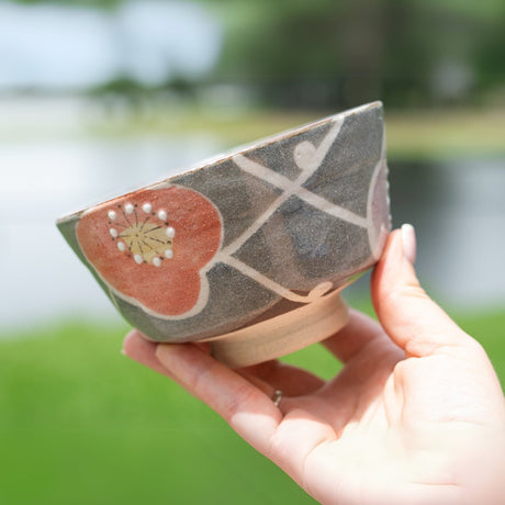 A hand holds the Gray with Flowers Ceramic Matcha Bowl from the Organic Japanese Matcha Set, set against a blurred outdoor background.